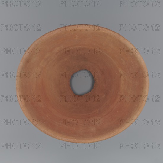 Cake baking mold. A vessel so-called flower pot for baking bread with a conical shape and a flat bottom. Shape irregular; straight, flaring spout; irregular bottom with a hole c. 6 cm diameter made before firing. Wheel-made from Nile clay of type NB2 Vienna System. Surface level; outer part near bottom and bottom finished with a knife or similar tool. Vessel color yellowish-red. Very well preserved. Rim diameter 22.3–25.2 cm; bottom diameter 10.2–11 cm; height 17.4 cm. c. 1550–1069 BC, New Kingdom, Deir el-Medina., A round, shallow object with a central irregular hole sits against a smooth background; the object displays a range of earthy terracotta and brown tones with subtle variations and darker spots, and the background is a solid cool gray.