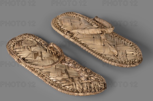 Sandals, footwear, grave goods, pair of papyrus sandals made using the ribbon technique. Edges finished with a cross-ribbed technique. c. 1550–c. 1069 BC; New Kingdom, Egypt, clothing, height 24.4 cm, width 11 cm, A pair of woven sandals made from natural fibers are shown on a solid gray background; the sandals display a braided and interlaced pattern with visible texture and fraying at the edges, and each sandal has a woven strap across the top and a toe post constructed from wrapped fibers. The colors visible include beige, tan, light brown, and darker brown variations in the fibers, with the background in medium gray.