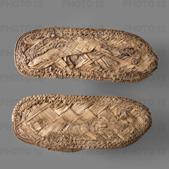 Sandals, pair of papyrus sandals made using the ribbon technique; edges finished with a cross-ribbed technique. Footwear, grave goods. c. 1550–c. 1069 BC New Kingdom, Egypt. Height 24.4 cm, width 11 cm., A pair of woven straw sandals viewed from the soles, showing a textured pattern of braided and interlaced straw fibers in various shades of tan and beige, with some darker brown strands and worn, frayed edges; the sandals rest on a smooth gray background.