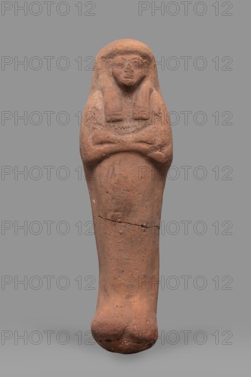 Ushabti;, Mummy-shaped funerary figurine, arms crossed on the chest, no tools. Smooth tripartite wig; very large eyes, wide nose, slightly twisted mouth. Inscription illegible. New Kingdom, 19th–20th Dynasty, ca. 1295–1069 BC. Ushabti; height 20 cm, width 6 cm., A single upright terracotta figure with a rounded head and stylized facial features, arms crossed over the chest, and a long tapering body ending in a rounded base, displayed against a flat gray background; the figure shows an overall reddish-brown clay color with variations of darker brown and small pale flecks, and surface irregularities and a visible horizontal crack across the lower torso.