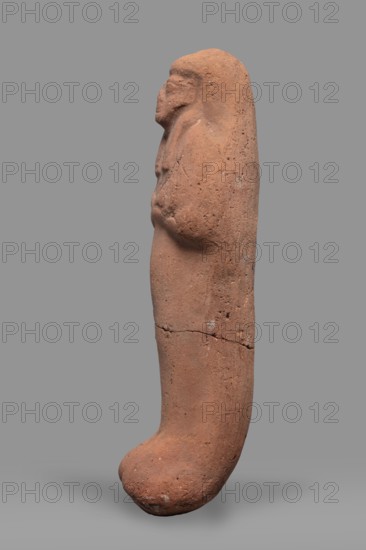 Ushabti. Funerary mummy-shaped figurine, arms crossed on the chest, no tools. On the head is a smooth tripartite wig. Very large eyes, a wide nose, a slightly twisted mouth. Inscription illegible. Ca. 1295–1069 BC; New Kingdom, 19th–20th Dynasty, Egypt. Ushabti; height 20 cm, width 6 cm., A tall, elongated reddish-brown clay figure shown in profile against a neutral gray background, with a rounded top and a curved, bulbous lower end, a few worn facial features and a suggestion of arms folded across the chest, a rough, pitted surface texture with visible cracks and darker spots, and overall earthy red and brown tones with some subtle lighter and darker variations.