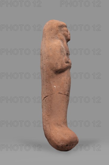 Ushabti, Funerary figurine mummy-shaped ushabti. Arms crossed on the chest, no tools. Smooth tripartite wig; very large eyes, wide nose, slightly twisted mouth. Inscription illegible. ca. 1295–1069 BC; New Kingdom, 19th–20th Dynasty, Egypt. Height 20 cm; width 6 cm., A weathered terracotta figurine in shades of reddish-brown and ochre stands upright against a plain gray background, showing a simplified human-like profile with a rounded head, a subtle nose and chin, and an elongated body tapering into a curved, bulbous lower end; the surface has a rough, pitted texture with small dark speckles and lighter patches, visible cracks and a horizontal seam-like break around the midsection, and faint contouring that suggests folded arms or raised hands across the chest.