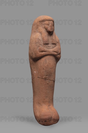Ushabti, Mummy-shaped funerary figurine, arms crossed on the chest, no tools. Smooth tripartite wig; very large eyes, wide nose, slightly twisted mouth. Inscription illegible. Ca. 1295–1069 BC; New Kingdom, 19th–20th Dynasty, Egypt. Ushabti; height 20 cm, width 6 cm., A single terracotta-colored figurine shaped like a wrapped human form stands against a plain gray background, showing a rounded head with a stylized face and a simple headdress, crossed arms folded over the chest, and a long narrow body that tapers into a bulbous foot-like base; the surface is rough and weathered with visible cracks and small dark spots, and the colors visible are terracotta, brown, gray, and small areas of darker blackish discoloration.