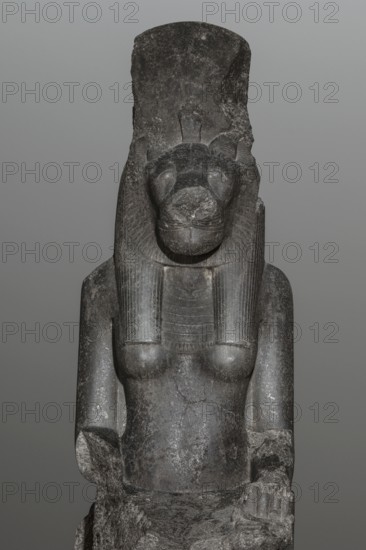 Statue of the goddess Sekhmet. Woman with a lioness's head seated on a throne, dressed in a long, close-fitting veil. Hands placed on her knees; in her left hand the goddess holds the ankh sign of life. Solar discs and uraei present. At the front of the throne, next to the goddess's legs, a column of hieroglyphs on each side. The sides of the throne are carved with a sema-taui motif. Egyptian deity; lioness-headed goddess. Sculpture, c. 1390–1352 BC, New Kingdom, 18th Dynasty reign of Amenhotep III, found at Karnak. Height 240 cm, width 61 cm, depth 123 cm, weight 2300 kg., A dark gray stone statue of a seated figure with a feline head and a tall, flat headdress, showing carved facial features and a stylized mane. The figure has a human torso with visible breasts and detailed, ribbed collar elements extending over the shoulders and chest. The arms rest at the sides, with one hand partially visible and resting on a rough, broken base. The surface of the statue shows variations in tone w...