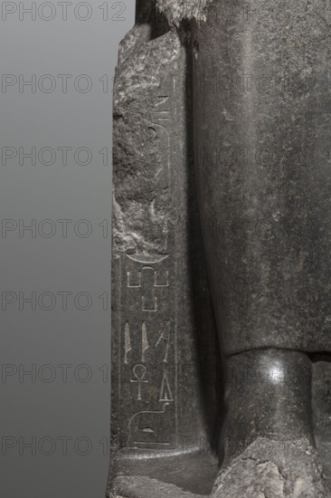 Statue of the goddess Sekhmet, Sekhmet mythology, solar discs, uraei, hieroglyphs, thrones, lionesses, Egyptian deities. Woman with a lioness's head seated on a throne, dressed in a long, close-fitting veil. Hands placed on her knees; in her left hand the goddess holds the sign of life. At the front of the throne, next to the goddess's legs, a column of hieroglyphs on each side. On the sides of the throne is carved a sema taui motif. c. 1390–1352 BC; New Kingdom, 18th Dynasty reign of Amenhotep III, found at Karnak. Sculpture; height 240 cm, width 61 cm, depth 123 cm, weight 2300 kg., A close-up view of a dark gray stone sculpture showing a section of a carved figure seated on a base, with a flat vertical slab beside the figure. The stone surface displays a range of gray tones from light to dark, with rough weathered textures and smoother polished areas. The seated figure's knee and part of the lower leg are visible, rounded and smoother with subtle highlights, while the adjacent slab ...