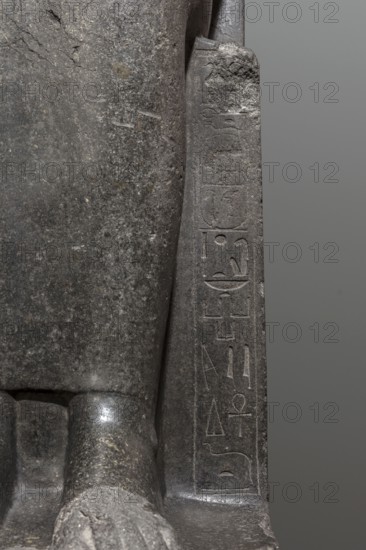 Statue of the goddess Sekhmet, Sekhmet mythology. Woman with a lioness's head seated on a throne, dressed in a long, close-fitting veil. Hands on her knees; in her left hand she holds the ankh sign of life. At the front of the throne, next to the goddess's legs, a column of hieroglyphs on each side. Sides of the throne carved with a sema-taui motif; solar discs and uraei present. c. 1390–1352 BC, New Kingdom, 18th Dynasty reign of Amenhotep III, found at Karnak. Sculpture; height 240 cm, width 61 cm, depth 123 cm, weight 2300 kg., A close-up view of a stone sculpture showing a smooth dark gray surface with lighter gray speckling and textured wear, a vertical carved panel with shallow incised markings, a rounded protruding element at the base, and a neutral medium gray background; visible colors include dark gray, light gray, and medium gray.