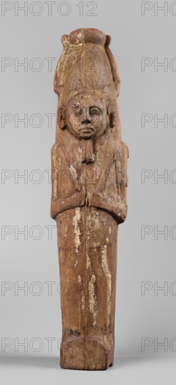 Papyrus container - figure of the god Ptah-Sokar-Osiris; atef crowns; Ptah-Sokar-Osiris mythology; atef crown with narrow feathers on a tripartite wig. The face is oval, wide; straight long nose; large mouth with fleshy lips; large ears; straight beard. Arms modeled, folded on the chest holding two nechacha whips. Torso wrapped in a shroud. Under the feet a large oval hole for inserting the papyrus plugged. On the back of the legs, a fragment of an inscription painted in black, yellow, and red on a white background. Type II E according to Raven; ca. 1295–ca. 945 BC; 19th–21st Dynasty, Egypt. Sculpture; material: papyri; height 60 cm; width 13.5 cm; depth 14 cm; weight 1.736 kg., A tall carved wooden figure with a weathered surface stands against a plain light gray background; the figure has a rounded headdress and a stylized face with closed or partially closed eyes and a neutral mouth, with hands held together at chest level and elongated, simplified arms and legs. The wood shows area...