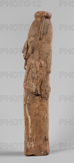 Papyrus container in the form of the god Ptah-Sokar-Osiris, Atef crown with narrow feathers on a tripartite wig. Oval, wide face with a straight long nose, large mouth with fleshy lips, large ears, and a straight beard. Modeled arms folded on the chest holding two nechacha whips. Torso wrapped in a shroud. Large oval hole under the feet for inserting papyrus plugged. Fragment of an inscription on the back of the legs painted in black, yellow, and red on a white background. Type II E according to Raven, ca. 1295–ca. 945 BC; 19th–21st Dynasty, Egypt. Sculpture; materials: papyri/wood papyrus container. Dimensions: height 60 cm, width 13.5 cm, depth 14 cm; weight 1.736 kg., A tall carved wooden figure shown in profile against a pale gray background, the wood displaying a range of brown tones from light tan to deeper brown with some pale worn areas and darker spots; the figure has a rounded top and an elongated body with a carved head in profile, a prominent nose and chin, and simplified a...