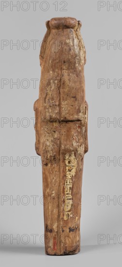 Papyrus container — figure of the god Ptah-Sokar-Osiris. Atef crown with narrow feathers on a tripartite wig. Oval, wide face with a straight long nose, large mouth with fleshy lips, large ears, and a straight beard. Modeled arms folded on the chest holding two nechacha whips. Torso wrapped in a shroud. Large oval hole under the feet for inserting papyrus plugged. Fragment of an inscription in black, yellow, and red paint on a white background on the back of the legs. Type II E according to Raven, ca. 1295–ca. 945 BC; 19th–21st Dynasty, Egypt. Sculpture papyrus container. Height 60 cm, width 13.5 cm, depth 14 cm, weight 1.736 kg., A vertical wooden figure with a weathered, rough surface in shades of brown and tan stands against a plain light gray background; the figure has a rounded top and an elongated body with visible grain, cracks, and areas of lighter beige peeling or flaked material, and a few small darker brown and black spots and streaks across its surface, with subtle hints of...
