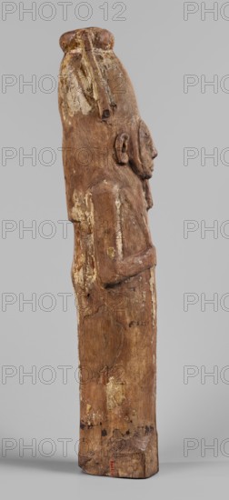 Papyrus container in the form of the god Ptah-Sokar-Osiris, Atef crown with narrow feathers on a tripartite wig. Oval, wide face with straight long nose; large mouth with fleshy lips, large ears, and a straight beard. Arms modeled and folded on the chest, holding two nechacha whips. Torso wrapped in a shroud. Large oval hole under the feet for inserting papyrus plugged. Fragment of an inscription on the back of the legs painted in black, yellow, and red on a white background. Type II E according to Raven, ca. 1295–ca. 945 BC; 19th–21st Dynasty, Egypt. Sculpture; material: papyri; height 60 cm, width 13.5 cm, depth 14 cm, weight 1.736 kg., A carved wooden figure shown in profile with a tall rounded headpiece and a pronounced nose and chin, the surface worn and textured with areas of flaking and abrasion; the figure's ear and folded arm are visible, and the carving stands on a rectangular base; colors visible include various shades of brown in the wood, patches of lighter beige where the...
