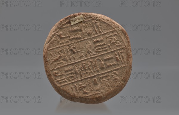 Mentuemhat's funeral cone, Mentuemhat ca. 700–650 BC. Clay cone with hieroglyphs; on the base a hieroglyphic inscription arranged in four lines. Inscriptions include name Nesiptah. Late Period, Western Thebes grave 34 on Asasipha, cone grave. Height 8.5 cm, width 12 cm, weight 0.628 kg. Davies-Macadam, Corpus No. 486, 664–332 BC., A round, flat object with a rough, textured surface in shades of brown and tan, showing a pattern of incised lines and shallow carved shapes arranged in horizontal bands across its face; the edges are irregular and slightly worn, and there is a small rectangular piece of material affixed near the top edge; the background is a smooth, neutral gray.
