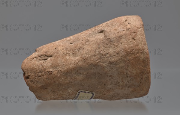 Mentuemhat's funeral cone;, Mentuemhat ca. 700–650 BCE. Hieroglyphs; Nesiptah ca. 700–650 BCE. On the base a hieroglyphic inscription arranged in four lines. Davies-Macadam, Corpus No. 486. Date: 664–332 BCE, Late Period. Western Thebes grave 34 on Asasipha, cone grave. Height 8.5 cm, width 12 cm, weight 0.628 kg., The image shows a rough, irregularly shaped object with a textured surface that appears worn and porous, displaying a range of brown tones from light tan to deeper reddish-brown with hints of gray; the object rests against a smooth background that is mainly gray, and a small rectangular patch with a patterned border is visible on the object's lower edge.