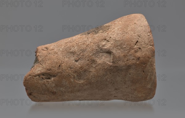 Mentuemhat's funeral cone, Mentuemhat ca. 700–650 BC, hieroglyphs, Nesiptah ca. 700–650 BC. On the base a hieroglyphic inscription arranged in four lines. Davies-Macadam, Corpus No. 486; 664–332 BC; Late Period, Western Thebes grave 34 on Asasipha, cone grave; height 8.5 cm, width 12 cm, weight 0.628 kg., A roughly conical, irregular object with a coarse, weathered surface fills the frame, showing a texture of pitted and cracked areas and small indentations. The object displays a range of earthy colors including reddish-brown, tan, and grayish-brown tones, with some darker brown spots and lighter beige highlights. The background is a smooth, neutral gray that contrasts with the object's rough surface. The object's profile includes a blunt end and a tapered end, and there are visible rough edges and small cavities on its surface.