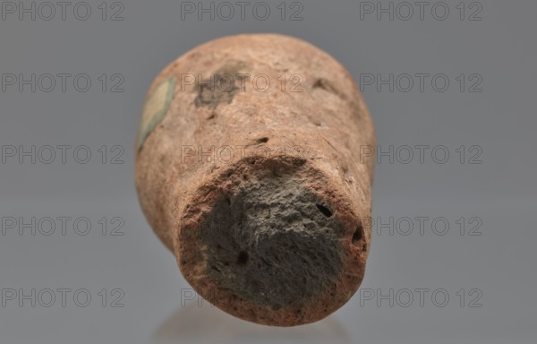 Mentuemhat's funeral cone, Mentuemhat ca. 700–650 BCE, hieroglyphs, Nesiptah ca. 700–650 BCE. On the base a hieroglyphic inscription arranged in four lines. Davies-Macadam, Corpus No. 486, 664–332 BCE; Late Period, Western Thebes grave 34 on Asasipha, cone grave. Height 8.5 cm, width 12 cm, weight 0.628 kg., A close-up view of a roughly cylindrical, weathered object with a textured, porous surface in shades of brown, reddish-brown, gray, and dark gray, shown against a smooth neutral gray background; the object's end facing the viewer appears rounded with irregular holes and rough, uneven edges, while the sides display worn, mottled patches and a small area of faded greenish color.