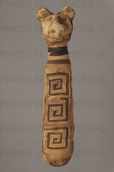 Cat mummy, Cat mummy decorated with a meander made of white and brown bandages. 30 BC–395 AD, Roman period, Egypt. Mummy; materials: fabrics, linen. Height 27.3 cm, width 6.5 cm, depth 6 cm; subjects: cats, meander ornament., A long fabric object shaped like a narrow body with a rounded head that has two rounded ears, wrapped in patterned strips of cloth showing a geometric spiral motif, with visible wear and fraying; the colors visible are tan, brown, dark brown, and black against a plain gray background.