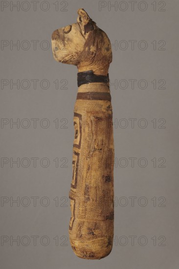 Cat mummy, unknown author; Cat mummy decorated with a meander made of white and brown bandages; 30 BC–395 AD, Roman period, Egypt; mummy; materials: fabrics, linen; dimensions: height 27.3 cm, width 6.5 cm, depth 6 cm; subjects: cats, meander ornament., A vertically oriented wrapped figure shaped like a long cylindrical form with a rounded base and a head-like top, covered in layered textile bands showing a worn, frayed texture; the textiles appear in shades of brown, tan, and dark brown with patterned darker bands and geometric markings visible on the body; a dark band encircles the neck area; the head-like top has folds and protrusions suggesting wrapped fabric and shows similar brown and tan tones; the background is a uniform neutral gray.