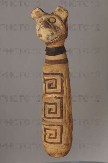 Cat mummy, cat mummy decorated with a meander made of white and brown bandages. 30 BC–395 AD, Roman period, Egypt. Materials: fabrics, linen. Type: mummy. Dimensions: height 27.3 cm, width 6.5 cm, depth 6 cm., A fabric object shaped like a long narrow cylinder with a rounded end and a simple head at the top, the head has two small rounded ears and stitched circular features for eyes and a protruding snout, the surface of the fabric shows worn and patched areas and rough texture, the body is wrapped in alternating horizontal bands and square spiral designs, the visible colors are tan, brown, dark brown, beige, and a muted gray background.