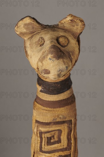 Cat mummy, Cat mummy decorated with a meander made of white and brown bandages. Materials: fabrics, linen. Subject: cats; ornament: meander. Date: 30 BC–395 AD, Roman period, Egypt. Type: mummy. Dimensions: height 27.3 cm, width 6.5 cm, depth 6 cm., A worn stuffed figure resembling an animal head and elongated body made of rough woven fabric in shades of tan, beige, brown, and dark brown, with rounded ears, a protruding snout, stitched circular eye shapes, a few darker spots and stains on the face, a banded neck with alternating dark and tan rings, and a patterned rectangular spiral design on the torso, all set against a plain gray background.