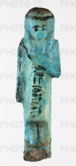 Shabti overseer Amenemone, A shabti figurine depicted in living attire, wearing a knee-length loincloth. In her left hand, bent at the elbow and folded across her chest, she holds a hoe placed on her right shoulder; the hoe's blade reaches down to her hand. The right arm hangs along the body. The face is small, triangular, with only the eyes clearly defined; other facial details are blurred. On the head is a three-part smooth wig tied with a headband, the ends of which fall down the back; the ears are uncovered. A column of hieroglyphic text on the headband at the front. Text, hoe and headband, and facial details painted in black. Circa 1069–c.945 BC, Third Intermediate Period, 21st Dynasty, Egypt. Shabti; height 8 cm, width 2.3 cm., A small statuette shaped like a standing human figure with a rounded head and stylized facial features, rendered in a turquoise-blue material with areas of darker blue and greenish patina, showing patches of black pigment on the hairline, eyes, and parts o...