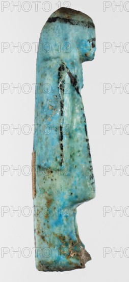 Shabti overseer Amenemone, A shabti funerary figurine depicted in living attire, wearing a knee-length loincloth. In her left hand, bent at the elbow and folded across her chest, she holds a hoe placed on her right shoulder; the hoe's blade reaches down to her hand. The right arm hangs along the body. The face is small and triangular, with only the eyes clearly defined; other facial details are blurred. On the head is a three-part smooth wig tied with a headband, the ends falling down the back and leaving the ears uncovered. A column of hieroglyphic text on the headband at the front. Text, hoe and headband, and facial details painted in black paint. Circa 1069–945 BC, Third Intermediate Period, 21st Dynasty, Egypt. Shabti; height 8 cm, width 2.3 cm., A small carved figurine photographed from the side showing a standing human-like form with a rounded head and a long straight body, with a subtle arm and leg profile visible; the surface shows a dominant turquoise-blue color with areas of ...