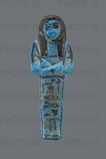 Ushabti Henut-netjeru. Mummy-shaped figurine shabti. Arms crossed on the chest, right folded over left, holding hoes in both hands; rectangular basket on the back; painted tools. Wearing a usech necklace on the chest and wide bracelets on the wrists painted jewelry. Long, smooth, three-part wig covering the ears; round face with large eyes; nose and mouth barely visible; wig and facial details painted. Painted inscription in one column on the front of the legs reading Shd Osiris Henut-netjeru, justified by the voice. Datation: ca. 1069–ca. 945 BC, Third Intermediate Period, 21st Dynasty, Egypt. Dimensions: height 9 cm, width 3 cm, depth 1.5 cm. Reference: Hnwt-ntrw -Ranke, Personennamen I, 243.25., A small blue and dark gray figurine shaped like a standing human figure with crossed arms, showing a rounded head with long dark hair and facial features, a tapered lower body with darker markings, and a textured surface with worn areas and speckles of blue and gray.