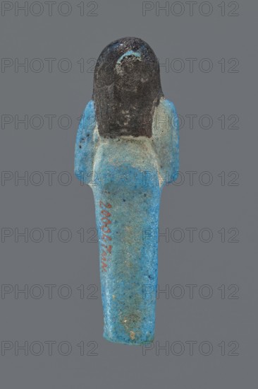 Ushabti Henut-netjeru. Mummy-shaped figurine shabti, height 9 cm, width 3 cm, depth 1.5 cm. Third Intermediate Period, 21st Dynasty, ca. 1069–ca. 945 BC. Arms crossed on the chest, right over left, holding hoes in both hands; a rectangular basket on the back with painted tools. Wearing a usech necklace and wide bracelets painted jewelry. Long, smooth three-part wig covering the ears; round face with large eyes; wig and facial details painted. Painted inscription in one column on the front of the legs reading: Shd Osiris Henut-netjeru, justified by the voice., The image shows a small human-shaped figurine viewed from the front against a plain gray background, with a rounded dark head and a long tapered body in bright blue with areas of lighter blue and speckled darker spots; the shoulders flare slightly and the surface appears textured and worn with patches of discoloration and roughness.