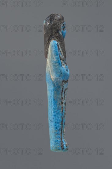 Ushabti Henut-netjeru, Henut-netjeru private name. Mummy-shaped figurine; shabti. Arms crossed on the chest, right over left, holding hoes in both hands; a rectangular basket on the back; painted tools. Wearing a usekh necklace and wide bracelets—painted jewelry. Long, smooth, three-part wig covering the ears; round face with large eyes, nose and mouth minimally modeled; wig and facial details painted. Painted inscription in one column on the front of the legs reading: Shd Osiris Henut-netjeru, justified by the voice. Date ca. 1069–ca. 945 BC, Third Intermediate Period, 21st Dynasty, Egypt. Dimensions: height 9 cm, width 3 cm, depth 1.5 cm., A small sculpted figure viewed in profile showing a standing person with long hair, arms folded against the body, and a long robe; the surface is predominantly bright blue with areas of darker blue and turquoise mottling, and the hair and some detailing appear dark gray to black, with a muted gray background.
