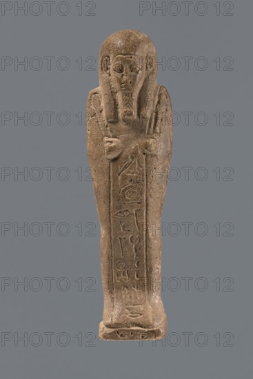 Ushabti, Mummy-shaped figurine, standing on a base with a pilaster at the back. Arms crossed on the chest, right folded over left; only the hands visible from under the shroud. Holds a hoe in the right hand and a pickaxe in the left; a twisted rope in the right hand supports a basket slung over the left shoulder. Engraved tools. Wears a smooth, three-part wig, high-domed; ears uncovered. Small face with large almond-shaped eyes, worn nose, mouth in a slight grimace. Long beard, slightly tapering, curled and braided at the end. Inscribed on the front of the legs from below the crossed arms to the toes, inscription column bordered on both sides by vertical lines. Date: 332–30 BC, Ptolemaic Period, Egypt. Type: shabti. Dimensions: height 10.5 cm, width 3.9 cm., A small carved figure in shades of brown with a rounded head and stylized facial features, long hair or a headdress framing the face, arms folded across the chest, a tapered body standing on a small rectangular base, with shallow i...