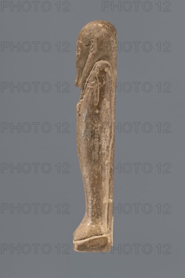Ushabti, Mummy-shaped figurine standing on a base with a pilaster at the back. Arms crossed on the chest, right folded over left; only the hands visible from under the shroud. Right hand holds a hoe and a twisted rope with a basket slung over the left shoulder; left hand holds a pickaxe. Engraved tools. Smooth three-part wig, high-domed with uncovered ears. Small face, large almond-shaped eyes, worn nose, mouth in a slight grimace. Long beard, slightly tapering, curled at the end and braided. Inscribed on the front of the legs from below the crossed arms to the toes, column of text bordered by vertical lines. 332–30 BC; Ptolemaic Period, Egypt. Shabti; height 10.5 cm, width 3.9 cm., A carved figure shown in profile against a solid gray background, rendered in a weathered brown-beige stone with lighter and darker patches, depicting a standing human-like form with a rounded head, a long beard or chin feature, a suggestion of facial features and an ear, elongated torso and legs, arms held...