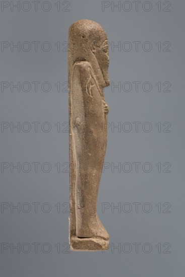 Ushabti, mummy-shaped figurine, standing on a base with a pilaster at the back. Arms crossed on the chest right folded over left; only the hands visible from under the shroud. Right hand holds a hoe and twisted rope with a basket slung over the left shoulder; left hand holds a pickaxe. Engraved tools. Smooth three-part wig, high-domed; ears uncovered. Small face with large almond-shaped eyes, worn nose, mouth in a slight grimace. Long beard, slightly tapering, curled and braided at the end. Inscribed on the front of the legs, from just below the crossed arms to the toes, column of text bordered by vertical lines. Date: 332–30 BC, Ptolemaic Period, Egypt. Object type: shabti. Dimensions: height 10.5 cm, width 3.9 cm., A slender upright stone figure photographed in profile against a neutral gray background, showing a carved human form with a rounded head, a long straight back, and a draped garment; the surface is textured and weathered with an overall brown and tan coloration, and there ...