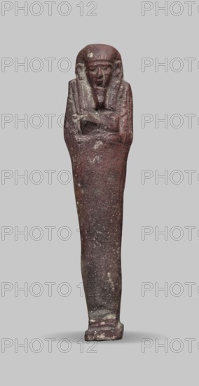 Ushabti, A shabti figurine dressed in a shroud stands on a base. On the head is a smooth wig falling in two strands on the chest. The false beard is smooth and straight. In his hands, crossed on the chest and protruding from under the shroud, he holds hoes in the shape of the letter A. 664–332 BC; Late Period 27th–30th Dynasty, Egypt. Ushabti; height 11.8 cm, width 2.8 cm., A single elongated statuette standing upright against a plain neutral-gray background, appearing as a human figure with crossed arms and a stylized head covering, rendered in a dark reddish-brown hue with patches of lighter, worn areas and speckled pale spots, showing a textured, weathered surface and a narrow rectangular base, with visible contours of a face and draped elements on the upper torso.