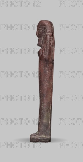 Ushabti, A shabti figurine dressed in a shroud stands on a base. On the head is a smooth wig falling in two strands on the chest. The false beard is smooth and straight. In his hands, crossed on the chest and protruding from under the shroud, he holds hoes in the shape of the letter A. 664–332 BC; Late Period 27–30th Dynasty, Egypt. Ushabti; height 11.8 cm, width 2.8 cm., A slender, upright carved figure shown in profile standing on a rectangular base, with a long, smooth body tapering slightly toward the top and a detailed head with a pronounced nose and long hair or headdress that falls down the back; the surface appears rough and worn with textured pitting and irregularities. The visible colors include reddish-brown, dark brown, and patches of grayish tones, with lighter gray around the base and faint darker spots along the shaft. The background is a uniform pale gray.