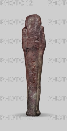 Ushabti, A shabti figurine dressed in a shroud stands on a base. On the head is a smooth wig falling in two strands on the chest. The false beard is smooth and straight. In his hands, crossed on the chest and protruding from under the shroud, he holds hoes in the shape of the letter A. 664–332 BC; Late Period 27th–30th Dynasty, Egypt. Ushabti, height 11.8 cm, width 2.8 cm., A vertically oriented slender statuette standing on a small base against a plain light gray background, with a rounded head and simplified body shape; the surface appears rough and weathered with visible texture variations and some cracks, and the overall color palette includes dark reddish-brown, muted brown, and areas of grayish-beige, with a few darker gray patches and subtle lighter speckles.