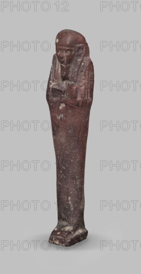 Ushabti, A shabti figurine dressed in a shroud stands on a base. On the head is a smooth wig, falling in two strands on the chest. The false beard is smooth and straight. In his hands, crossed on the chest and protruding from under the shroud, he holds hoes in the shape of the letter A. Late Period, 664–332 BC 27th–30th Dynasty, Egypt. Ushabti; height 11.8 cm, width 2.8 cm., A standing carved figure in a reddish-brown stone with worn surfaces, showing a stylized human head with a rounded top and detailed facial features, a narrow elongated body that tapers toward the base, and arms folded across the chest; the surface has areas of lighter discoloration and speckling, and the object casts a soft shadow on a plain light gray background.