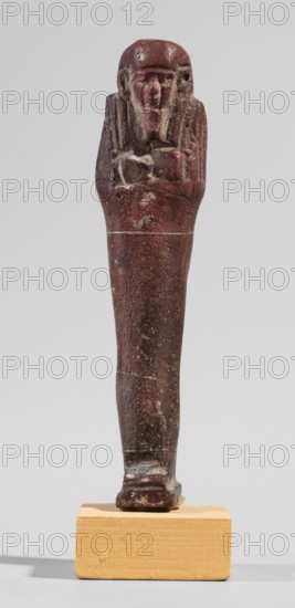 Ushabti, A shabti figurine dressed in a shroud stands on a base. On the head is a smooth wig, falling in two strands on the chest. The false beard is smooth and straight. In his hands, crossed on the chest and protruding from under the shroud, he holds hoes in the shape of the letter A. 664–332 BC; Late Period 27th–30th Dynasty, Egypt. Ushabti; height 11.8 cm, width 2.8 cm., A slender carved figurine in a deep reddish-brown color stands upright on a small rectangular pale yellowish-beige block; the figurine has a stylized human face and elongated head, with a few lighter worn patches and speckles of off-white and gray on its surface, a torso with arms crossed over the chest, a long tapered lower body, and a flattened foot resting on the block, all set against a plain light gray background.