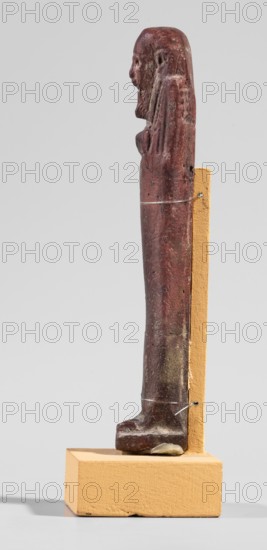Ushabti, A shabti figurine dressed in a shroud stands on a base. On the head is a smooth wig, falling in two strands on the chest. The false beard is smooth and straight. In his hands, crossed on the chest and protruding from under the shroud, he holds hoes in the shape of the letter A. 664–332 BC; Late Period 27–30th Dynasty, Egypt. Ushabti; height 11.8 cm, width 2.8 cm., A slender carved figure in reddish-brown stands upright on a rectangular ochre base, supported at the back by an ochre vertical block; the figure shows a carved head with facial features and a long headdress or hair, a narrow torso with faint arm details along the sides, and feet resting on the base, with visible surface texture, small pits, and areas of lighter wear revealing muted brown and grayish tones against the dominant red and ochre colors.