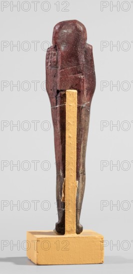 Ushabti, A shabti figurine dressed in a shroud stands on a base. On the head is a smooth wig, falling in two strands on the chest. The false beard is smooth and straight. In his hands, crossed on the chest and protruding from under the shroud, he holds hoes in the shape of the letter A. Late Period, 664–332 BC 27th–30th Dynasty, Egypt. Ushabti; height 11.8 cm, width 2.8 cm., A tall slender vertical figurine stands on a rectangular base; the figurine is primarily dark reddish brown with some darker brown and black areas and a few lighter worn spots, and it is supported by a vertical ochre-yellow strip attached to the base and fastened around the figurine with thin silvery wires; the base is ochre-yellow and sits on a neutral pale gray surface against a light gray background.