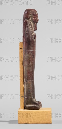 Ushabti, A shabti figurine dressed in a shroud stands on a base. On the head is a smooth wig, falling in two strands on the chest. The false beard is smooth and straight. In his hands, crossed on the chest and protruding from under the shroud, he holds hoes in the shape of the letter A. 664–332 BC; Late Period 27–30th Dynasty, Egypt. Ushabti; height 11.8 cm, width 2.8 cm., A vertical carved figure mounted on a tan rectangular base and supported by a tan vertical backboard, the figure is elongated and stylized with a rounded head, prominent nose and chin, and a suggestion of an arm and hand against the torso; the surface shows a weathered, pitted texture with patches of dark reddish-brown, deep brown, and areas of lighter brown and beige, as well as some muted greenish spots, and thin clear wires looped around the figure to hold it to the support.