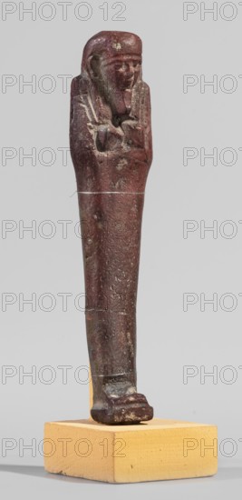 Ushabti, A shabti figurine dressed in a shroud stands on a base. On the head is a smooth wig falling in two strands on the chest. The false beard is smooth and straight. In his hands, crossed on the chest and protruding from under the shroud, he holds hoes in the shape of the letter A. 664–332 BC; Late Period 27th–30th Dynasty, Egypt. Ushabti; height 11.8 cm, width 2.8 cm., A small statuette of a standing figure with folded arms displayed on a rectangular block base; the figure has a rounded head with visible facial features and a headdress, and the body tapers down to a flat foot resting on the base; the surface shows an aged, rough texture with worn areas and small pits; colors visible include deep reddish-brown on the figure, lighter brown and pale tan on the base, and small areas of lighter discoloration on the figure.