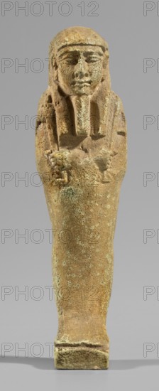 Ushabti, wig and beard, funerary figurine Mummy-shaped shabti standing on a base, supported at the back by a pillar. Arms crossed on the chest with only the hands protruding from under the shroud, placed at the same level slightly apart; hoes in both hands with slightly curled handle ends. In the right hand an additional twisted rope from which a basket is slung over the left shoulder. Slightly convex tools. Three-part smooth wig; long straight beard tapering to a braided end. Large ears, almond-shaped eyes, pronounced eyebrows, broad nose, full lips in a slight grimace. Not inscribed. Late Period, 664–332 BC 28th–30th Dynasty, Egypt. Shabti; height 11.7 cm, width 3.2 cm., A small carved figure with a rounded head and stylized facial features, a headdress with straight bangs and lappets framing the face, arms crossed over the chest holding small objects, and a tapered lower body standing on a rectangular base; the surface shows a weathered, pitted texture with areas of roughness. The v...