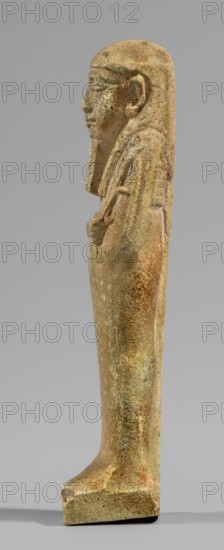 Ushabti, Restitution. Wigs, beards facial hair, funerary figurine mummy-shaped shabti standing on a base, supported at the back by a pillar/pilaster. Arms crossed on the chest with only the hands protruding from under the shroud, placed at the same level slightly apart. Hoes in both hands; the ends of the hoes' handles are slightly curled. In the right hand an additional twisted rope with a basket slung over the left shoulder. Slightly convex tools. On the head a three-part smooth wig. A long straight beard tapering toward the bottom, braided into a plait. Large ears, almond-shaped eyes, pronounced eyebrows, broad nose, full lips in a slight grimace. Not inscribed. 664–332 BC; Late Period 28th–30th Dynasty, Egypt. Height 11.7 cm, width 3.2 cm., A small standing statue of a person shown in profile against a plain gray background, rendered in a weathered beige and light brown material with mottled greenish and rusty brown spots, with a long head covering or hair that falls to the shoulde...