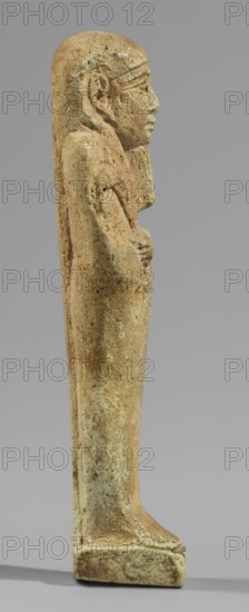 Ushabti, Mummy-shaped funerary figurine, standing on a base, supported at the back by a pillar/pilaster. Arms crossed on the chest with only the hands protruding from under the shroud, placed at the same level slightly apart; hoes in both hands with slightly curled ends of the handles. In the right hand an additional twisted rope with a basket slung over the left shoulder. Slightly convex tools. On the head a three-part smooth wig; long straight beard tapering to a braided tip. Large ears, almond-shaped eyes, pronounced eyebrows, broad nose, full lips in a slight grimace. Not inscribed. Late Period, 664–332 BC 28–30th Dynasty, Egypt. Shabti; height 11.7 cm, width 3.2 cm., A vertical stone figure in profile facing right, showing a carved head with a stylized wig and defined facial features, a torso with folded arms held against the chest, and a long straight lower body standing on a rectangular base; the surface shows a weathered, pitted texture with a mottled palette of beige, tan, lig...