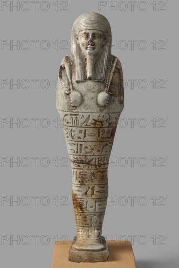 Ushabti of General Chaihorpata, Chaihorpata ca. 380–343 BC. Shabti, mummy-shaped figurine, standing on a base with a flat pillar at the back. Arms crossed on the chest with only the hands protruding from under the shroud; left hand holds a pickaxe, right hand a hoe, and a twisted string with a basket slung over the left shoulder. Convex tools. Wears a three-part wig styled in narrow strands with main strands ending in narrow borders; ears uncovered. Long, straight, narrow braided beard. Carefully modeled facial details with proportionate features; eyes and eyebrows marked with a soft line, mouth in a slight grimace; nose damaged. Inscription engraved and placed in nine rows separated by thin lines around the figurine's legs. 380–343 BC; Late Period, 30th Dynasty, Egypt. Height 20 cm; width 5.5 cm., A small carved figure stands upright on a rectangular base with a smooth rounded top, rendered in off-white and pale gray tones with areas of brownish staining and weathering; the figure has...