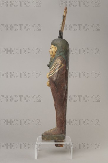 Figurine of Osiris in the form of a human mummy wearing a hemhem crown with ram's horns. On the chest is a usekh necklace with falcon heads at the ends. Clenched hands are placed on the chest, one above the other. Below the right hand begins a column of text with the name and filiation. At the back, from the edge of the wig to the feet, a pillar with a column of hieroglyphic inscriptions—the beginning of a hymn to Osiris, the heir of Atum. The figurine is painted cherry red; the visible parts of the body are yellow; the wig is navy blue; the necklace consists of white, red, and blue stripes. The background of the inscriptions is yellow. The figurine belongs to Isetemachbit, a musician of the god Amun-Re, daughter of the priest Mentuemhat; her mother was probably Takereret, known as Heres. Similar to type IV B according to Raven. 4th–3rd century BC 30th Dynasty–early Ptolemaic period, Egypt. Sculpture; height 54.5 cm, width 12.5 cm, depth 9.5 cm., A side view of a standing wooden figure...