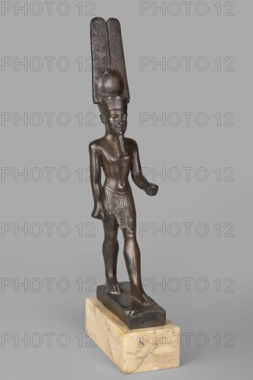 Figurine of the god Amun. Amun mythology. Figurine in a standing posture, wearing a helmet with a solar disc and feathers. Inlaid eyes. Right hand lowered with a clenched fist; left hand bent at the elbow with a clenched fist. Date with base: 664–525 BC; Late Period, 26th Dynasty, Egypt. Material: figurine. Weight 1.04 kg with base. Height 26 cm; width 3 cm; depth 8 cm., A small dark bronze figure of a standing male wearing a tall, elaborate headdress and a short skirt, mounted on a rectangular light beige marble base; the figure has a smooth, polished surface with visible sculpted details such as a beard, defined muscles, and textured skirt pleats, with the headdress showing vertical ridges and a rounded form, and the overall colors visible are dark brown bronze, lighter golden-brown highlights on the figure, and creamy beige with subtle veining on the marble base, set against a uniform light gray background.