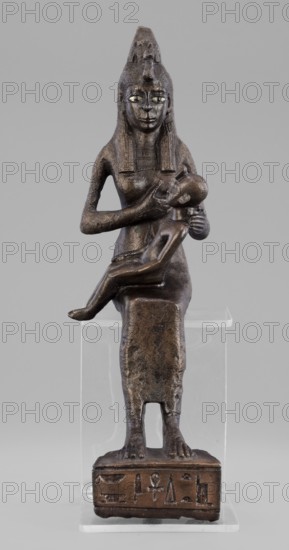Figurine of the goddess Isis nursing Horus, Isis mythology, Horus mythology, uraeus, hieroglyphs, goddesses, Egyptian deities, Egyptian art, Egyptian sculpture. The figurine depicts a seated Isis with the child Horus on her lap. Isis supports Horus with her left hand and offers him her breast with her right. Isis is dressed in a narrow, close-fitting robe. On her head is a fragment of the base of a crown. Horus is naked; his neck is adorned, and his head is adorned with a lock of youth hanging from the right side, with a uraeus placed above his forehead. On the base of the figurine is a hieroglyphic votive text. 525–343 BC; Late Period, 27th–30th Dynasty, Egypt. Sculpture; height 21.7 cm, width 7 cm, depth 10.5 cm, weight 1.22 kg., A dark brown metallic figurine of a seated female figure holding a smaller child on her lap is shown against a plain light gray background; the larger figure has a tall headdress, elongated facial features with inlaid lighter-colored eyes, long straight hair...