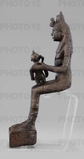 Figurine of the goddess Isis nursing Horus. The figurine depicts a seated Isis with the child Horus on her lap. Isis supports Horus with her left hand and offers him her breast with her right. Isis is dressed in a narrow, close-fitting robe. On her head is a fragment of the base of a crown. Horus is naked, wearing a neck ornament; his head bears a lock of youth hanging from the right side and a uraeus placed above his forehead. On the base of the figurine is a hieroglyphic votive text. Late Period, 525–343 BC 27th–30th Dynasty, Egypt. Sculpture. Height 21.7 cm; width 7 cm; depth 10.5 cm; weight 1.22 kg., A dark brown metallic sculpture of a seated adult figure in profile holding a smaller childlike figure on its knee, both figures showing textured surfaces and defined facial profiles, the adult wearing a tall headdress and a long garment draped along the back, the adult's hand resting on the child's shoulder while the child faces forward with a rounded head and small limbs, the adult s...