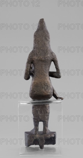Figurine of the goddess Isis nursing Horus, Isis mythology, Horus mythology, uraeus, hieroglyphs, goddesses, Egyptian deities, Egyptian art, Egyptian sculpture. The figurine depicts a seated Isis with the child Horus on her lap. Isis supports Horus with her left hand and offers him her breast with her right. Isis is dressed in a narrow, close-fitting robe. On her head is a fragment of the base of a crown. Horus is naked; he wears a neck ornament, and his head is adorned with a lock of youth hanging from the right side and a uraeus placed above his forehead. On the base of the figurine is a hieroglyphic votive text. 525–343 BC; Late Period, 27th–30th Dynasty, Egypt. Sculpture. Height 21.7 cm, width 7 cm, depth 10.5 cm, weight 1.22 kg., A small sculptural object shown from the back, made of rough textured dark brown metal with some lighter brown highlights, featuring a tall pointed head or hood and a torso with rounded shoulders and short bent arms; the figure sits on a narrow seat with ...