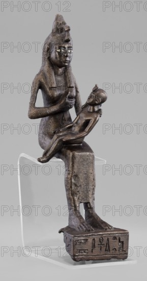 Figurine of the goddess Isis nursing Horus, Isis mythology, Horus mythology, uraeus, hieroglyphs, goddesses, Egyptian deities, Egyptian art, Egyptian sculpture. The figurine depicts a seated Isis with the child Horus on her lap. Isis supports Horus with her left hand and offers him her breast with her right. Isis is dressed in a narrow, close-fitting robe. On her head is a fragment of the base of a crown. Horus is naked; his neck is adorned, and his head bears a lock of youth hanging from the right side and a uraeus placed above his forehead. On the base of the figurine is a hieroglyphic votive text. 525–343 BC; Late Period, 27th–30th Dynasty, Egypt. Sculpture; height 21.7 cm, width 7 cm, depth 10.5 cm, weight 1.22 kg., A dark brown bronze statuette of a seated adult figure wearing a tall headdress and a patterned collar holds a smaller nude child figure on their lap; the adult has elongated hair or a wig falling to the shoulders and a calm facial expression with light-colored inlaid e...