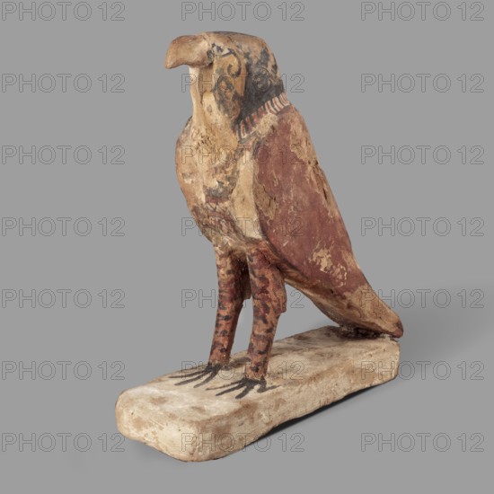Figurine of the falcon Sokaris, guardian of the necropolis; Horus mythology. A falcon stands on a thick rectangular base, resting its tail on the back edge. The bird's head is slightly raised; the beak long and curved. The body is long with wings slightly plastically defined in their upper part; the lower parts of the wings merge with the tail. The head is black on the sides and back; the front of the head is yellow with a black diagonal check. The eye is marked with white and black paint. A white stripe around the neck. The wings are black and red. Bird legs with red and black lines. Egypt, 664–30 BC Late Period–Ptolemaic Period. Sculpture; height 24.3 cm, width 20.5 cm., A painted bird-shaped sculpture perched on a rectangular base, with a curved beak and a rounded head, showing worn and chipped painted surfaces; the sculpture displays a mix of brown, reddish-brown, beige, black, and faded ochre tones, with darker patterned markings on the head and legs and black stylized talon shape...